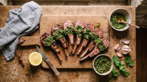 Grilled lamb cutlets fanned across a wooden serving board, deep char marks on pink medium-rare meat, generously spooned with a coarse green salsa verde flecked with parsley and capers.